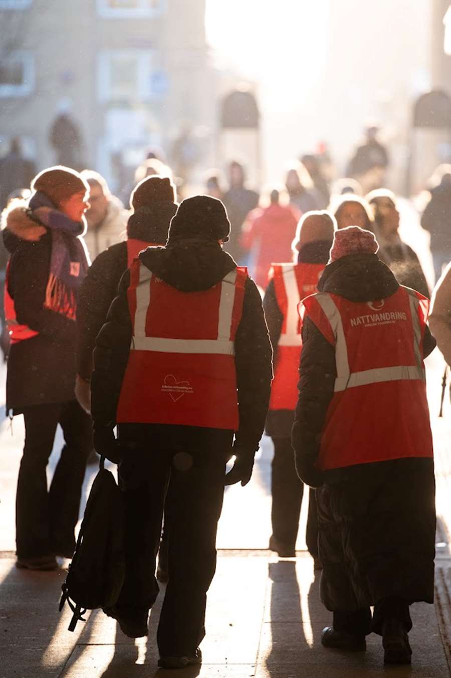 Ungdomsjouren är ute och går med volontärer. Solen skiner i motljus och personerna i bilden har röda västar från nattvandring.nu.