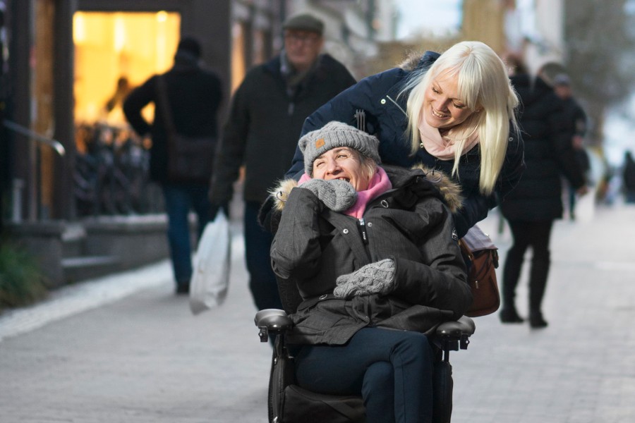 Två personer går på en gata. Den ena kör den andra i rullstol. Båda ser glada ut.