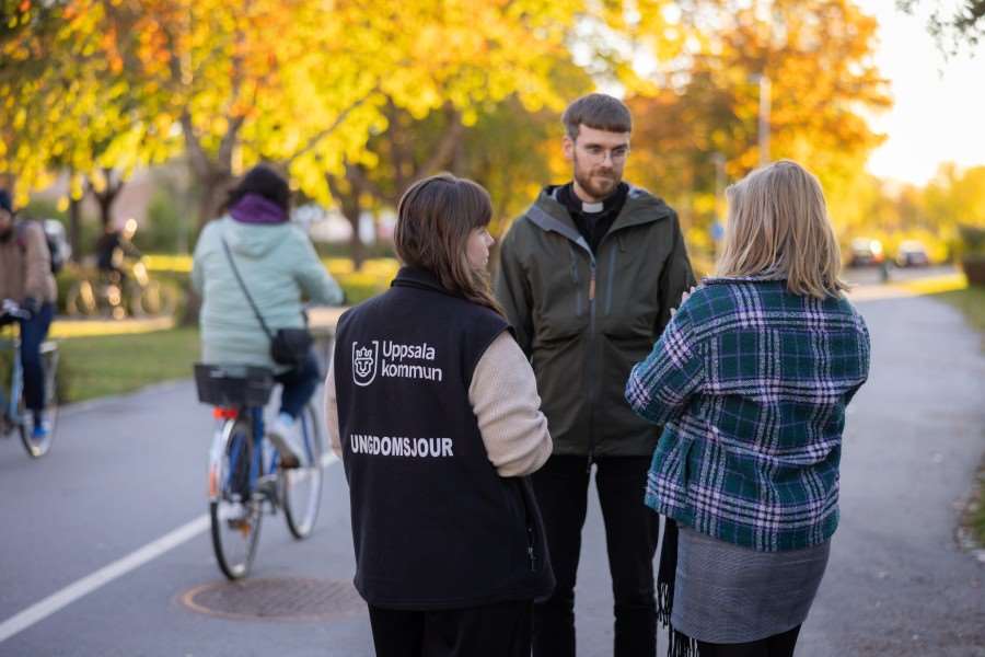 Tre personer står och pratar med varandra. En av personerna ha en jacka som det står Ungdomsjouren på. En av personerna har en prästkrage runt halsen.